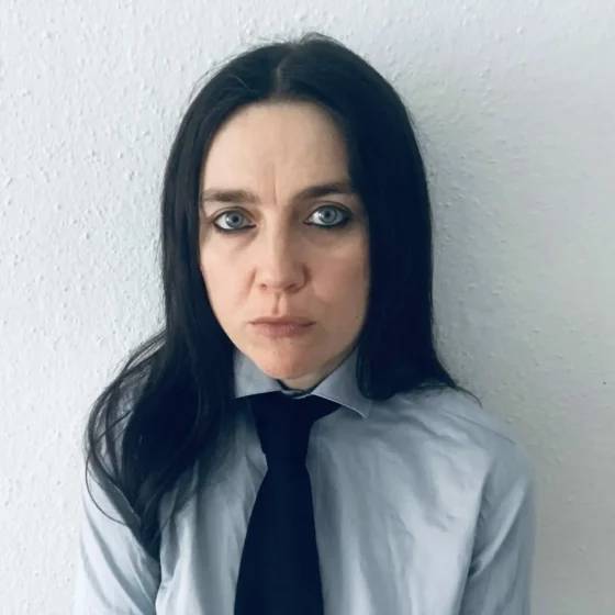 A woman stands in front of a gray wall and looks into the camera. Her long, straight black hair falls over her shoulders; she is wearing a gray shirt and a black tie. Her eyes are outlined in black with eyeliner.