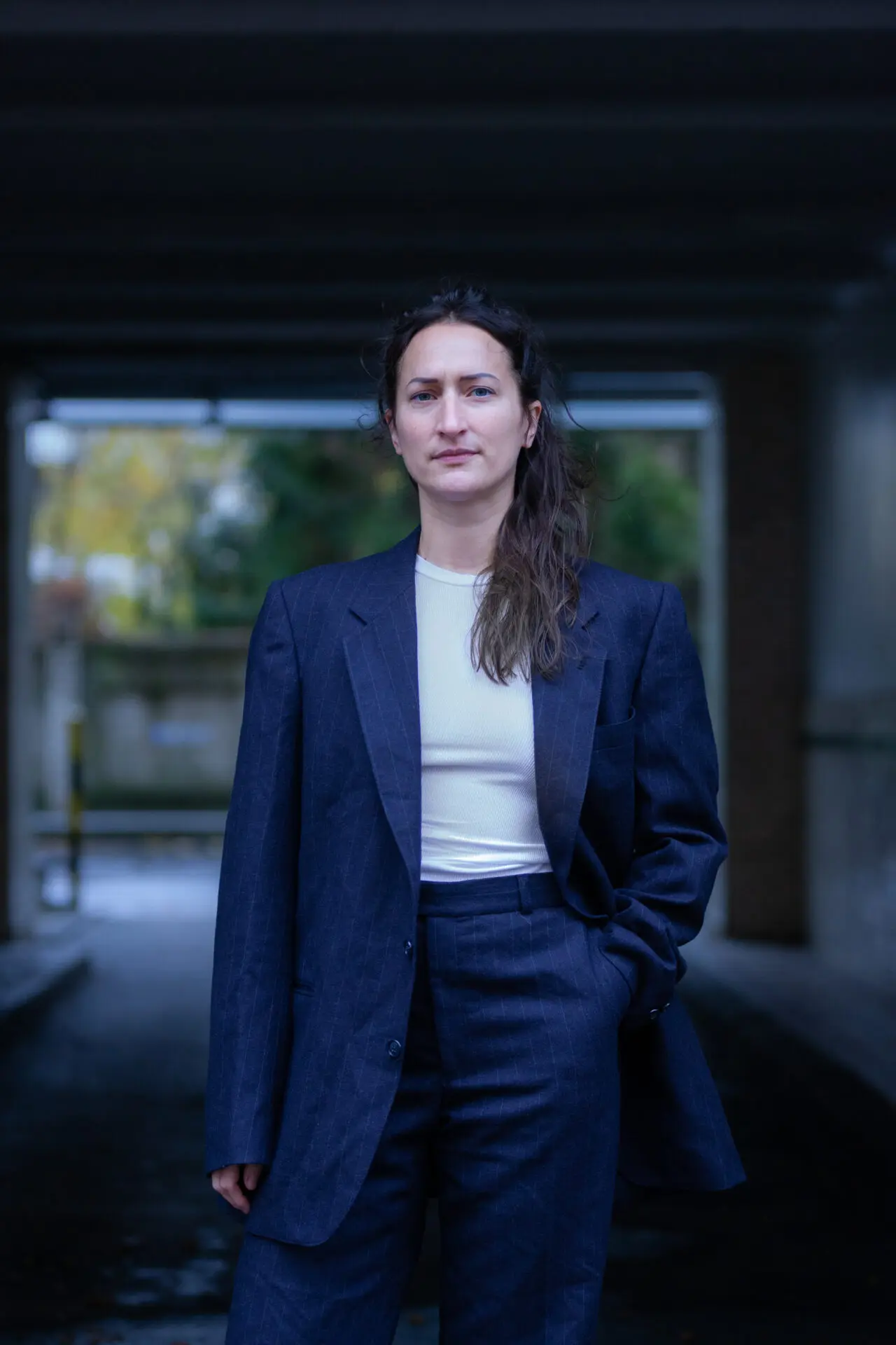 A person is standing in front of a doorway, the background in the picture is blurred. The person is white and is wearing dark blue trousers and a dark blue jacket over a white T-shirt. The left hand is tucked into the trouser pocket. The hair is dark brown, wavy and laid forward over the left shoulder. The person is looking into the camera.