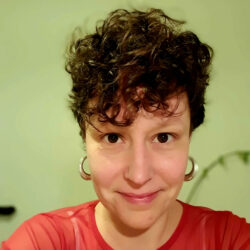 The image shows the face and upper torso of a white person with short brown curly hair, wearing a red mesh top and large silver hoop earrings. The person is looking directly at the camera with their brown eyes and is smiling. The background is blurry but you can sense that there is a plant behind them.