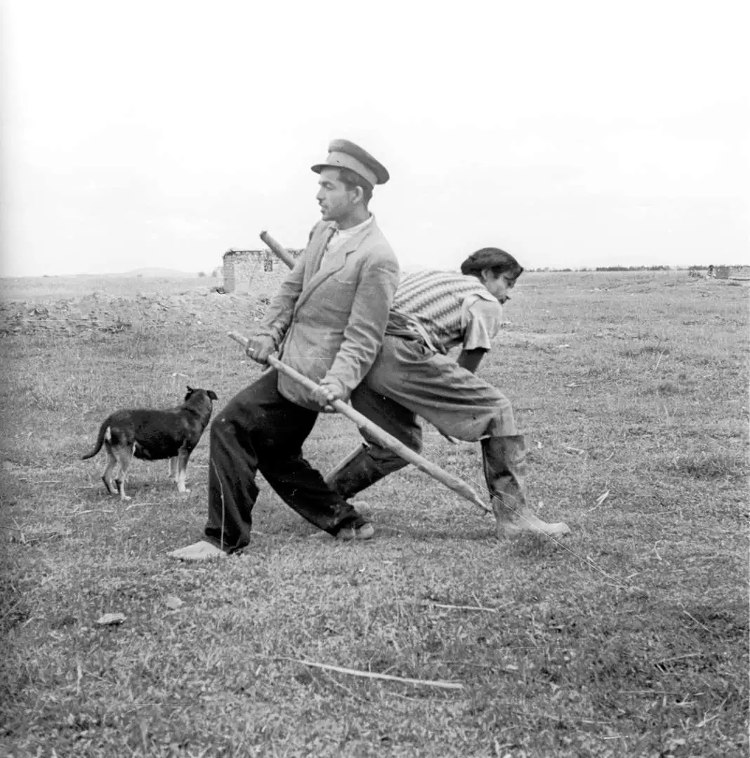 Black and white image. Two men are standing in a wide field, back to back, each holding a stick and dancing. A dog can be seen to their left and a wide horizon stretches out above their heads.