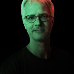The picture shows a man of about fifty with gray hair and a beard, wearing glasses and a black shirt. He looks into the camera with a slight smile. The background is dark and the man's face is illuminated by green and red lights, creating an interesting visual contrast.