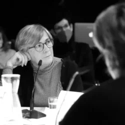 The black and white photo shows a portrait of a white woman with glasses sitting at a table in front of a microphone and listening to someone with interest. Her head is tilted to the side and resting on her hand. She is wearing a thin turtleneck and a light black cardigan. Her mottled gray hair is half-long. In front of her is her name badge and a glass of water. She is part of a discussion group. Some of the audience can be seen in the background.