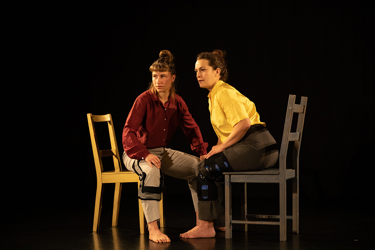 Two women are sitting on chairs and looking in the same direction. They are both wearing gray-brown pants, each with a splint on the knee, one is wearing a red shirt, the other a yellow one. Black molleton curtain in the background.
