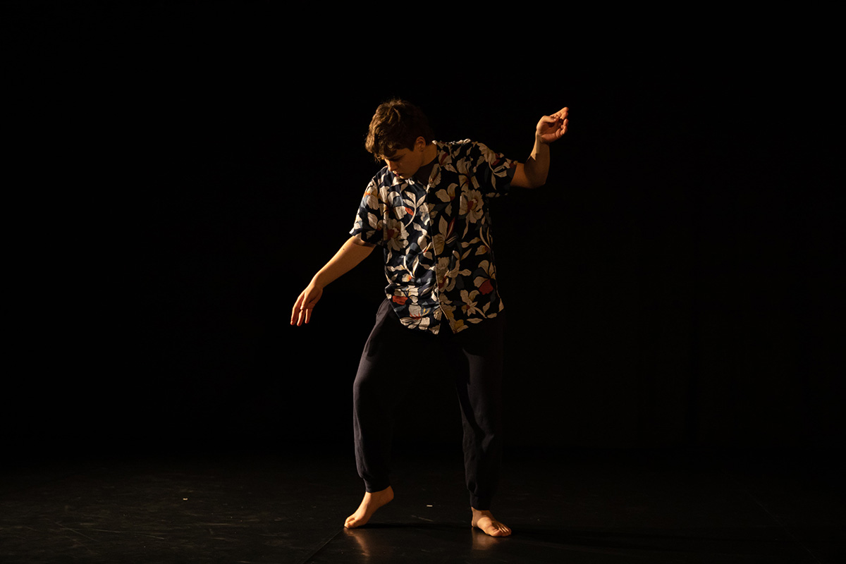 The picture shows a person in dance-like movement, barefoot, wearing black trousers and a colorful floral shirt. The gaze is lowered towards the ground, the right arm stretches into the air. Black background.