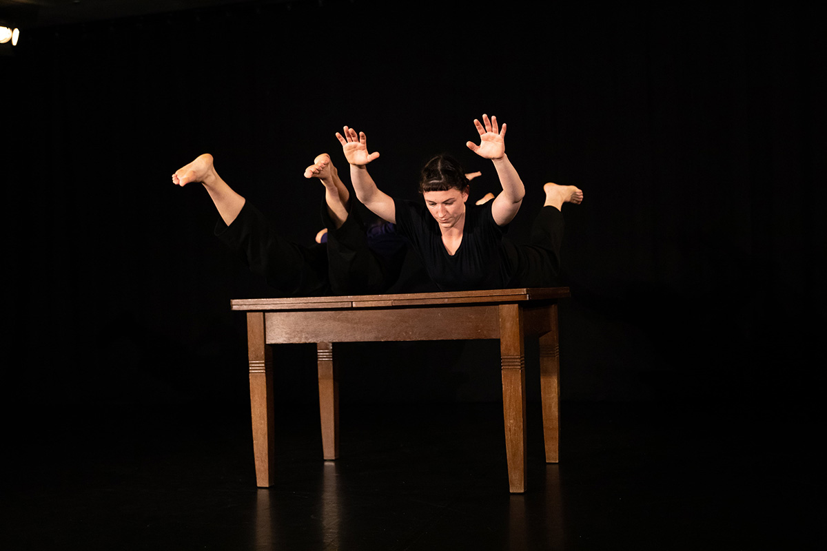 Two bodies are placed on a brown wooden table. On the right, a woman dressed in black with her stomach on the table, her arms and legs stretched upwards; on the left, a person with only her feet and legs visible. Black background.