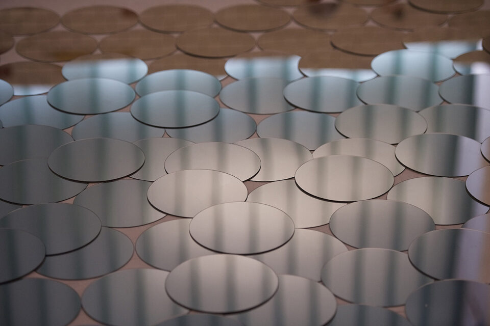 Das Bild zeigt einen hellen Boden der mit mehreren kreisrunden Spiegelplatten übersäht ist. Ein streifenförmiges Schattenmuster spiegelt sich darin.
