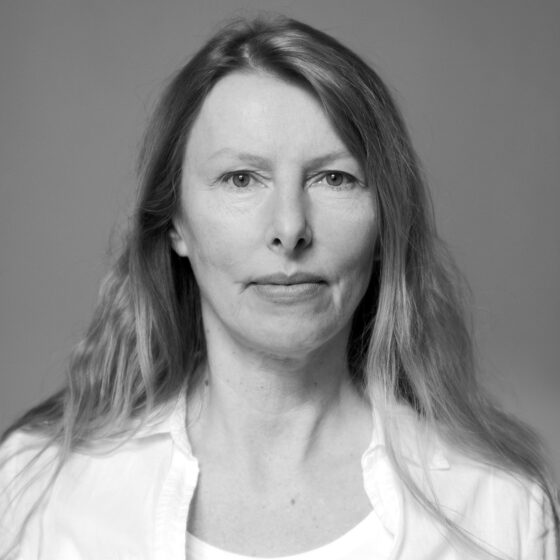 The black and white image shows a woman with long, straight blonde hair looking directly into the camera. She is wearing a wide white shirt with the top buttons undone and a white undershirt underneath. Her hair falls softly over her shoulders.