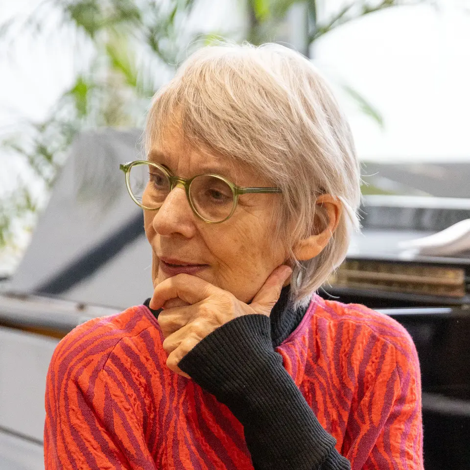 A woman in her 70s rests her chin on her left hand and tilts her head slightly to the right. Her hair is short and white-grey, she is wearing light green glasses and a red patterned top under a black long-sleeved top. The background is blurred, the outline of a green plant can be made out.