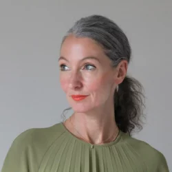 Isabel Raabe in front of a gray wall, her upper body leaning to the right, her face looking to the side. Her gray wavy hair is tied in a braid at the nape of her neck, she is wearing a muted green top, a gold necklace with a pendant, gold hoop earrings and bright orange lipstick.