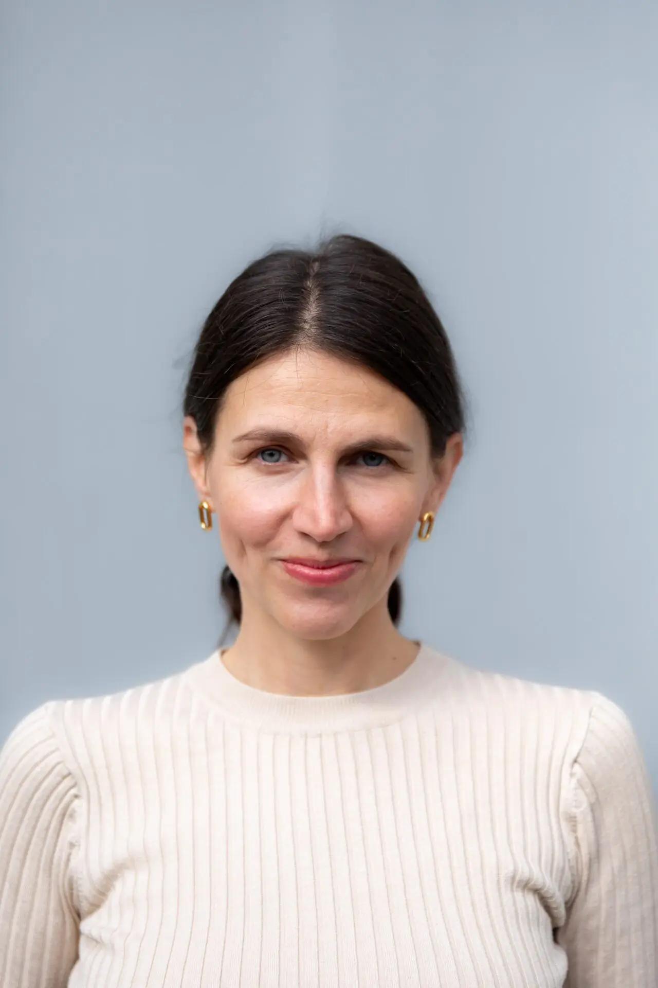 The picture shows a woman against a soft gray background, looking directly into the camera and smiling slightly with her mouth closed. Her dark brown straight hair is tied into a plait at the nape of her neck. She is wearing a beige ribbed long-sleeved top and gold hoop earrings