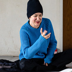Kirsten Maar is sitting on the floor and gesticulating with her right hand. She is wearing black trousers, a blue sweater and a black cap. An open notebook lies in front of her. The floor she is sitting on is gray, a whitish-beige wall can be seen in the background and a dark brown wooden beam on the right edge of the picture.