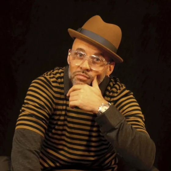The picture shows Storm, a middle-aged man with a well-groomed and graying five-day beard, a green Fedora hat on his head, large glasses with transparent frames on his nose and a silver hoop earring in his left ear. In this picture, he looks rather deliberate and thoughtful, which is partly due to his seated cross-legged pose, but also because he is holding his left hand on his chin. The background of the picture is completely black. This makes the light green of his hat and the thin stripes of his top stand out in contrast.