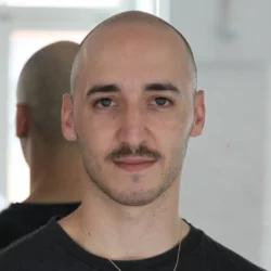 Nitsan Margaliot looks directly into the camera, wearing a black T-shirt, moustache, bald head and a chain with a gold pendant. He is standing in front of a mirror in which the outline of a window is reflected.