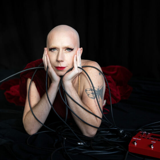 Olympia Bukkakis, a white drag queen with minimal makeup, a shaved head and long earrings in a long dress of crimson velvet lies among a tangled mess of cables. She looks into the camera with an expression of ironic vulnerability. The visual reference is that of a cheesy soprano publicity shot, but she means it.