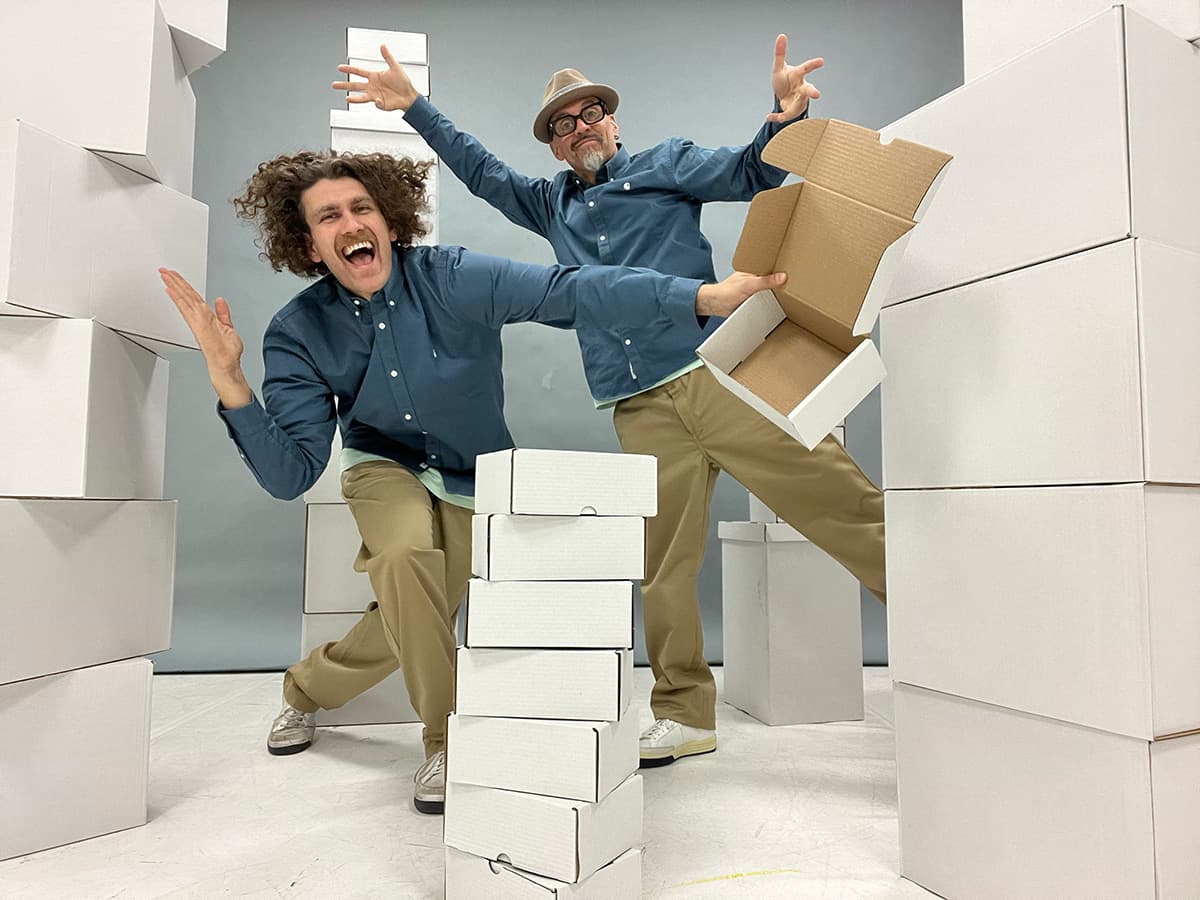 Two male figures are standing in the middle of several stacks of white cardboard boxes, both in representative dance poses and smiling at the camera. Both are wearing wide beige pants and blue denim shirts. The person at the back wears a beige hat and black glasses, the person in front has shoulder-length wavy brown hair.