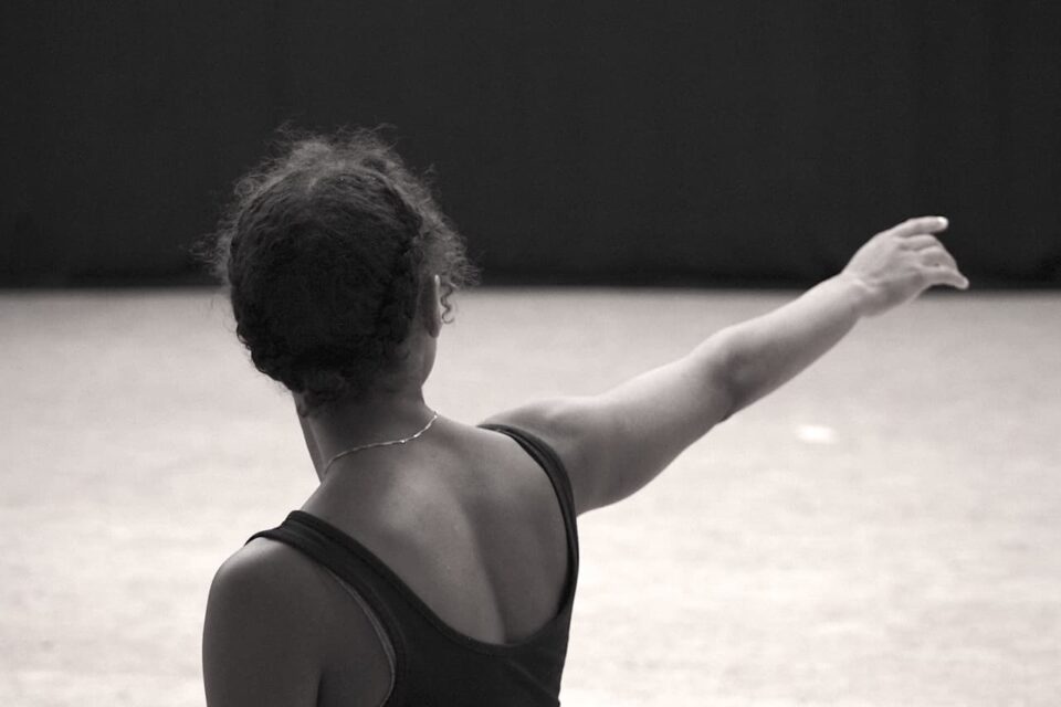 A black female person stands with her back turned to the camera in the center of the black-and-white image, pointing into the distance with her right arm outstretched. She is wearing a black sleeveless top and a fine link necklace.