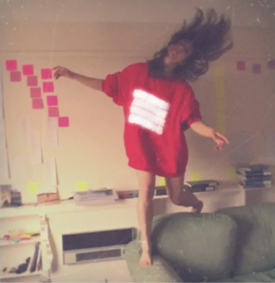 A dancer with long dark hair and wearing a red sweatshirt with blurry white lettering dances while standing precariously on the armrest of a sofa. In the background is a white walls with post-it notes and a thin ledge on which various books are stacked.