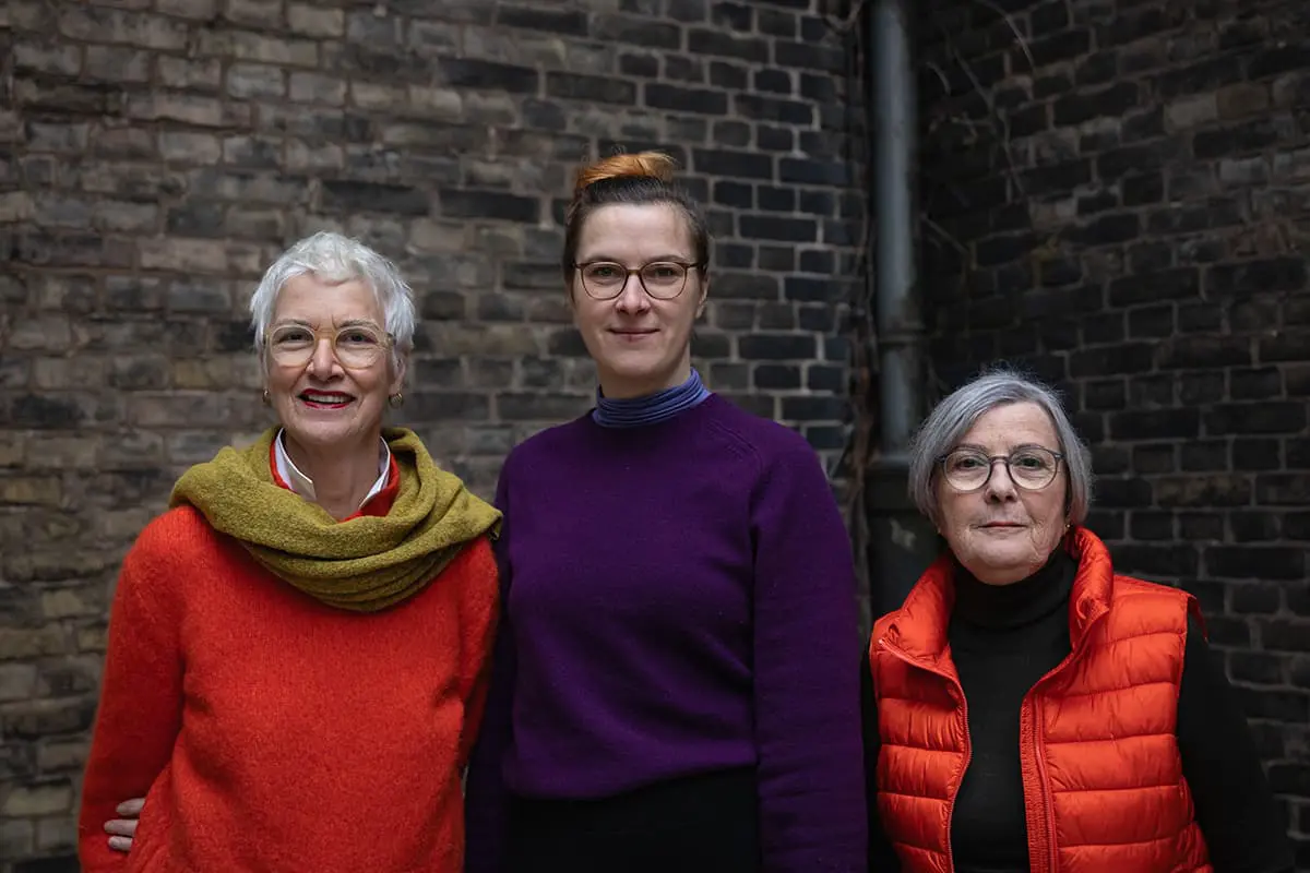 Three women stand next to each other in front of a dark brown brick wall. All three are looking into the camera. On the left, Claudia Feest with short white-grey hair, light-colored glasses, bright red wool sweater and mustard-yellow scarf. In the middle, Christine Henniger with auburn hair tied up in a high bun, light brown glasses and purple wool sweater. On the right, Claudia Henne with gray mid-length hair, gray-brown glasses, black turtleneck sweater and bright red quilted vest.