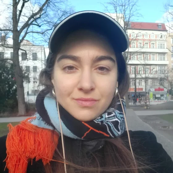The photo is a selfie of myself (Teresa Fazan). I am smiling, wearing a black cap, a black winter coat, and a black-blue-orange scarf around my neck. The sky is light blue, and I am standing in front of buildings' facades and some leafless trees.