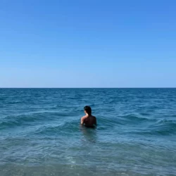 a person stands in a blue-green sea, turned away from the observer. the sky above the horizon is a clear spectrum of blue.