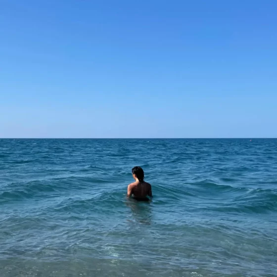a person stands in a blue-green sea, turned away from the observer. the sky above the horizon is a clear spectrum of blue.