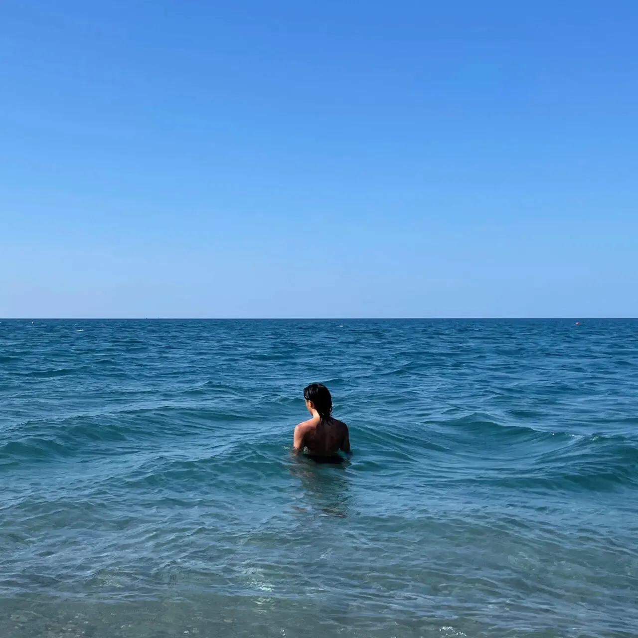 a person stands in a blue-green sea, turned away from the observer. the sky above the horizon is a clear spectrum of blue.