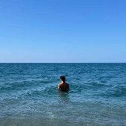 eine person steht in einem blaugrünen meer, vom betrachter abgewandt. der himmel über dem horizont ist ein klares blau-spektrum.