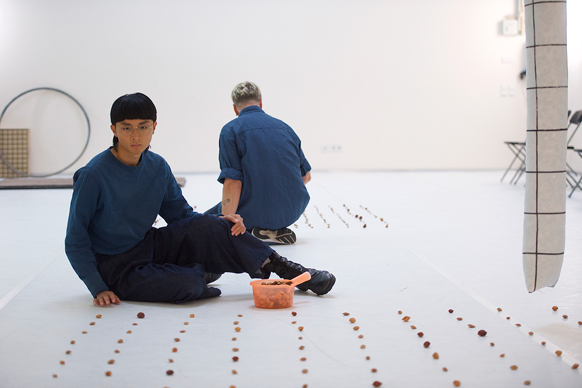 A person with black hair sits on the floor and looks into the camera; in front of her is a plastic bucket with fruit pits, with many geometrically arranged fruit pits scattered on the floor. The outline of another person can be seen in the background, also sitting on the floor with her back to the camera. The floor is white, the wall in the background is also white. A hula hoop can be seen leaning against the wall on the left edge of the picture.